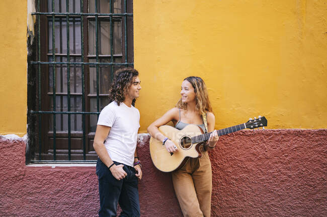 Sorridente giovane donna che guarda il fidanzato mentre suona la chitarra contro il muro, Santa Cruz, Siviglia, Spagna — Foto stock