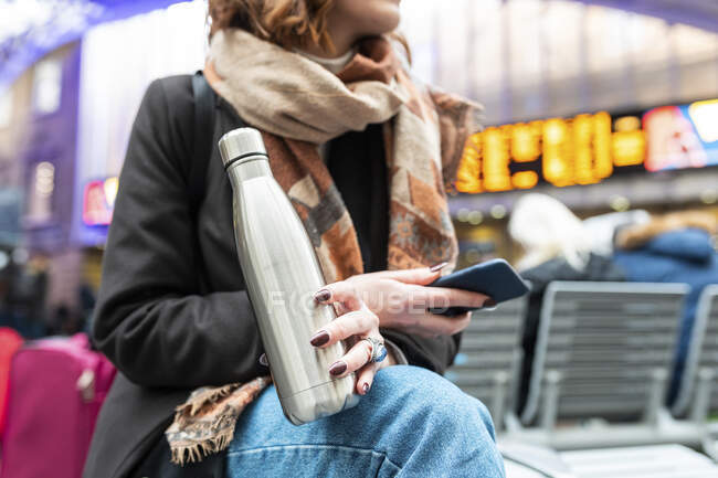 Жінка сидить на залізничній станції з термоподібною колбою та мобільним телефоном. — стокове фото