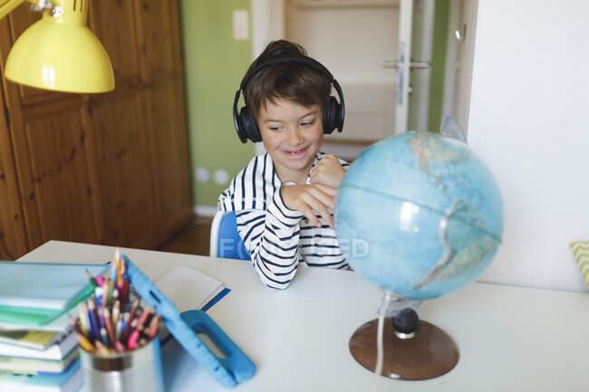 Menino fazendo homeschooling com um globo e usando tablet e fones de ouvido em casa — Fotografia de Stock