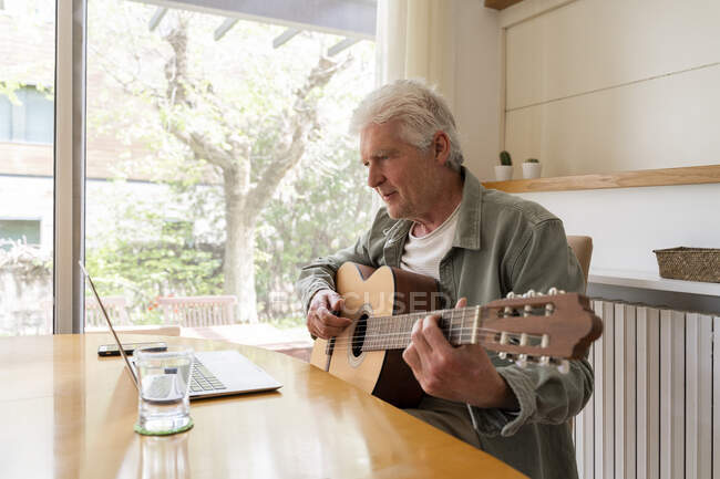 Pensionato uomo anziano imparare a suonare la chitarra attraverso tutorial online sul computer portatile a casa — Foto stock