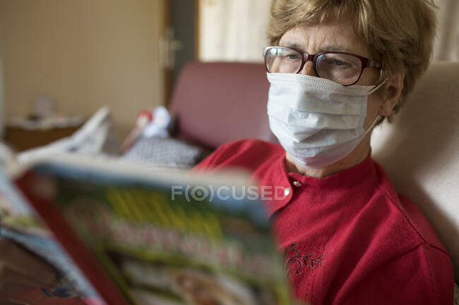 Старшая женщина читает Романа дома и носит защитную маску — стоковое фото
