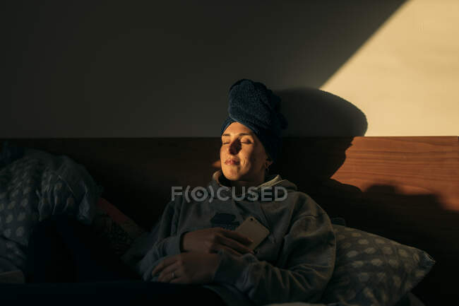 Jeune femme avec la tête enveloppée dans une serviette couchée dans le lit à la maison — Photo de stock