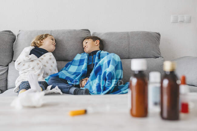 Irmãos doentes sentados no sofá em casa com medicamentos no tampo da mesa em primeiro plano — Fotografia de Stock