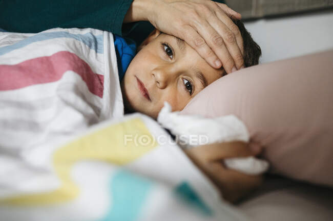 Portrait of sick boy lying in bed while his mother touching his forehead — Stock Photo
