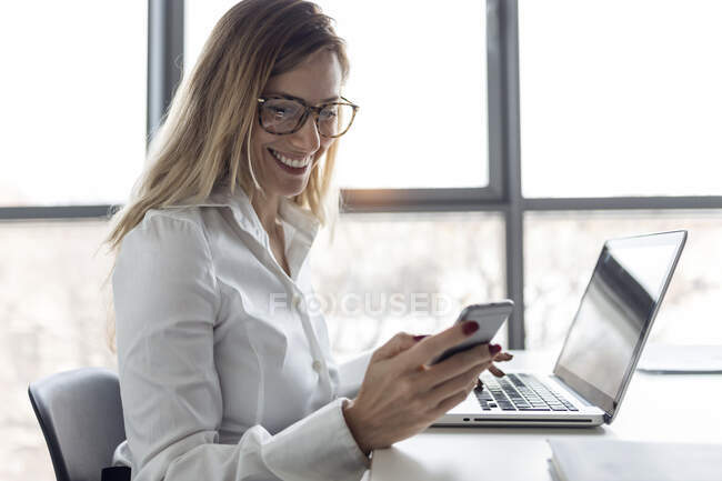 Деловая женщина, сидящая в офисе, работает на ноутбуке, используя смартфон — стоковое фото