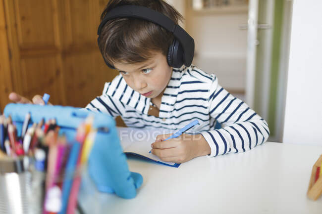 Хлопчик, який займається домашнім вивченням та записом у записнику, використовує вдома планшет та навушники. — стокове фото