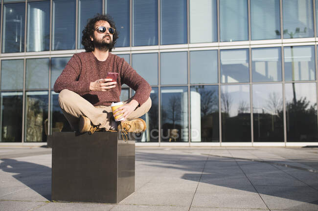 Портрет бородатого чоловіка, який сидить на відкритому повітрі з кавою, щоб піти і смартфон насолоджується сонячним світлом — стокове фото