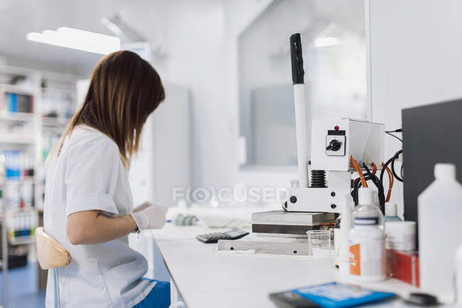 Vista lateral del médico femenino con cabello castaño largo sentado en el escritorio mientras trabaja en el laboratorio - foto de stock