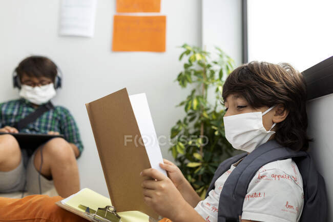 Niños con máscaras estudiando mientras están sentados a distancia en la escuela - foto de stock