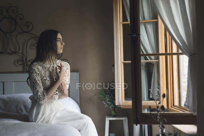 Giovane donna in elegante abito da sposa seduto sul letto guardando fuori dalla finestra — Foto stock