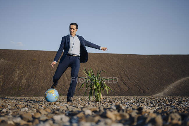 Homme d'affaires mature jouant au football avec un globe sur une pointe de mine désaffectée — Photo de stock