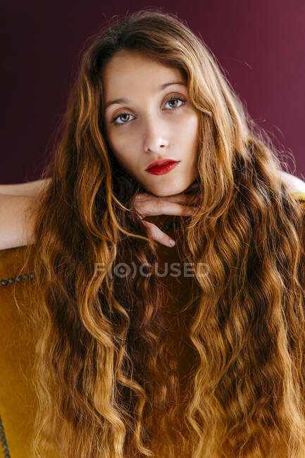 Portrait of young woman with long brown wavy hair leaning on chair against colored background — Stock Photo
