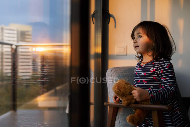 Portrait of little girl with teddy bear looking out of window at sunset — Stock Photo