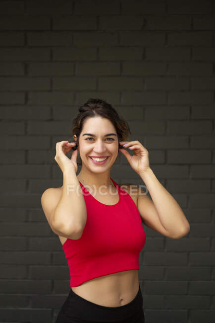 Smiling female athlete listening music through wireless headphones while standing against brick wall — Stock Photo