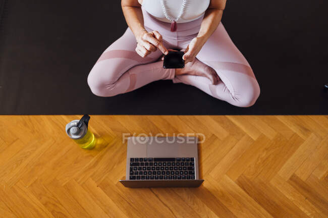 Frau benutzt Smartphone und Laptop, während sie zu Hause auf Turnmatte sitzt — Stockfoto