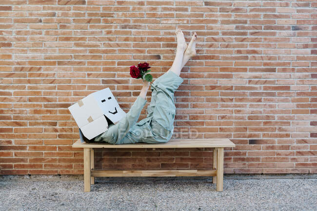 Woman holding red rose, wearing cardboard box with happy face, lying on bench in front of brick wall — Stock Photo