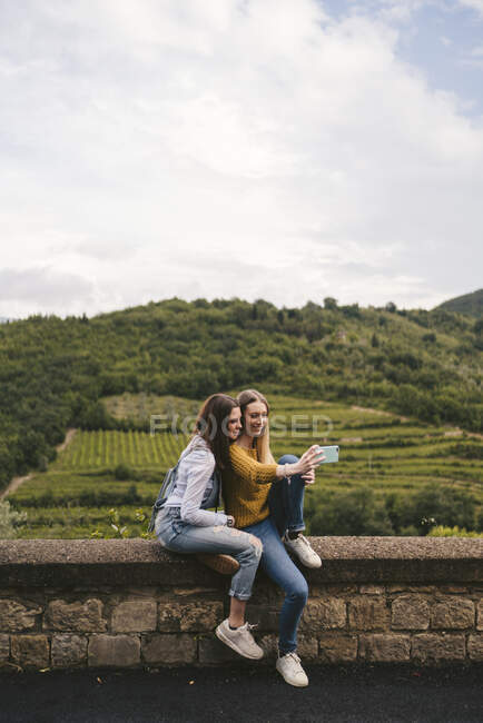 Две молодые женщины сидят на стене в сельской местности, делают селфи, Греве в Кьянти, Тоскана, Италия — стоковое фото
