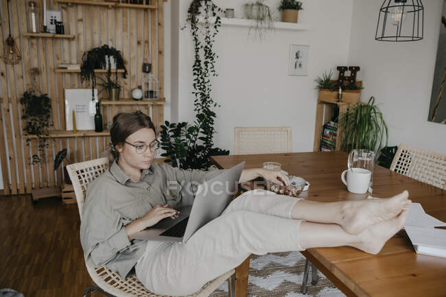 Mujer joven usando el ordenador portátil mientras está sentado en la mesa con los pies en la casa - foto de stock