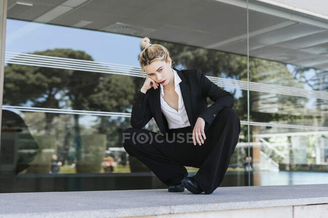 Portrait of young businesswoman wearing black pantsuit crouching in front of an office building — Stock Photo
