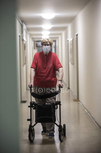 Femme âgée portant un masque et marchant avec un marcheur à roues dans le couloir de la maison de retraite — Photo de stock