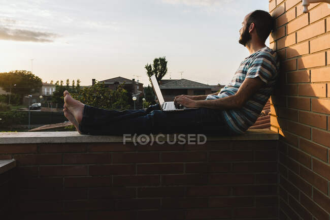 Uomo premuroso con computer portatile rilassante sul muro di contenimento contro il cielo durante il tramonto — Foto stock
