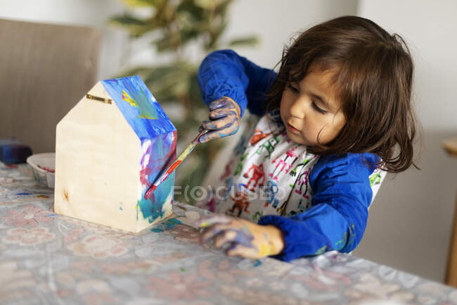 Linda chica pintando pequeña casa modelo de madera con pincel en la sala de estar - foto de stock