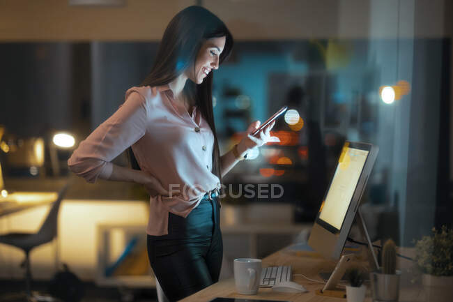 Felice giovane donna in piedi alla scrivania in ufficio guardando lo smartphone — Foto stock