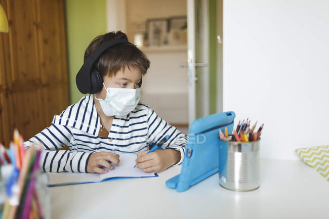 Junge macht Hausaufgaben und schreibt am Notizbuch, benutzt Tablet und Kopfhörer, trägt während der Coronakrise zu Hause eine Maske — Stockfoto
