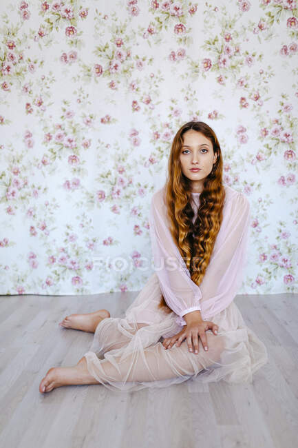 Портрет уверенной молодой женщины с длинными каштановыми волосами, сидящей на цветочных обоях — стоковое фото