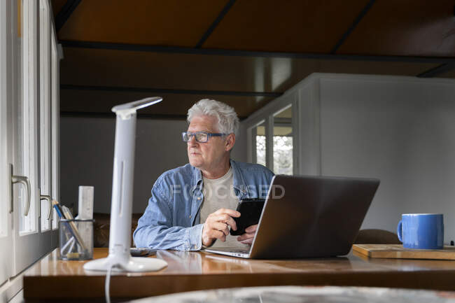 Pensativo homem idoso segurando telefone inteligente com laptop na mesa em casa — Fotografia de Stock
