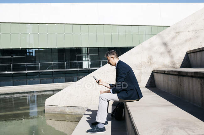 Homme d'affaires aux cheveux gris assis dans les escaliers d'une piscine à l'aide d'un téléphone portable — Photo de stock
