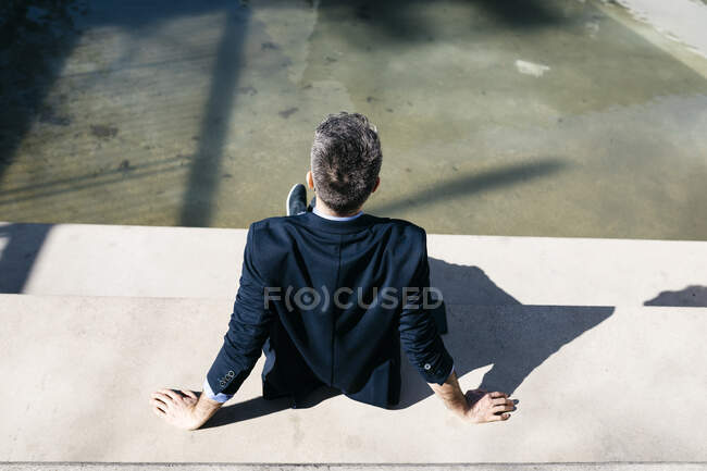 Vue arrière og homme d'affaires aux cheveux gris assis sur les escaliers d'une piscine — Photo de stock