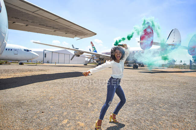 Веселая молодая женщина танцует с сигнальной ракетой на взлетно-посадочной полосе аэропорта в солнечный день — стоковое фото