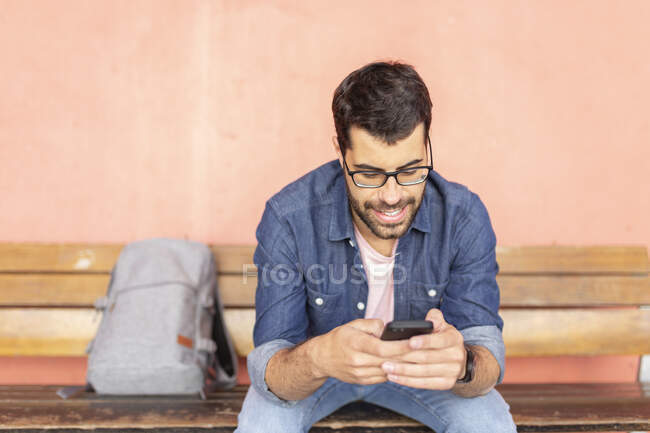 Retrato de homem sorridente sentado em banco de madeira usando smartphone — Fotografia de Stock