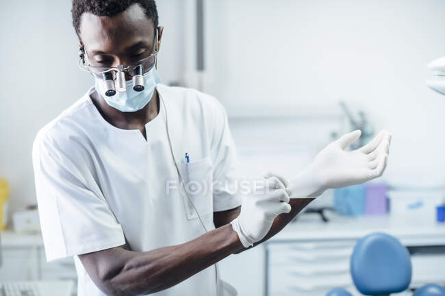 Retrato de dentista con lupa y máscara poniéndose guantes quirúrgicos en su práctica médica - foto de stock