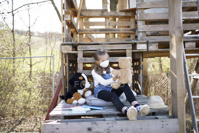 Mädchen mit Schutzmaske sitzt im Garten am Baumhaus und spielt während der Corona-Quarantäne allein mit ihren Kuscheltieren — Stockfoto