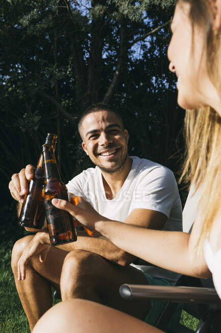 Smiling man toasting beer bottle with girlfriend while sitting at tourist resort — Stock Photo