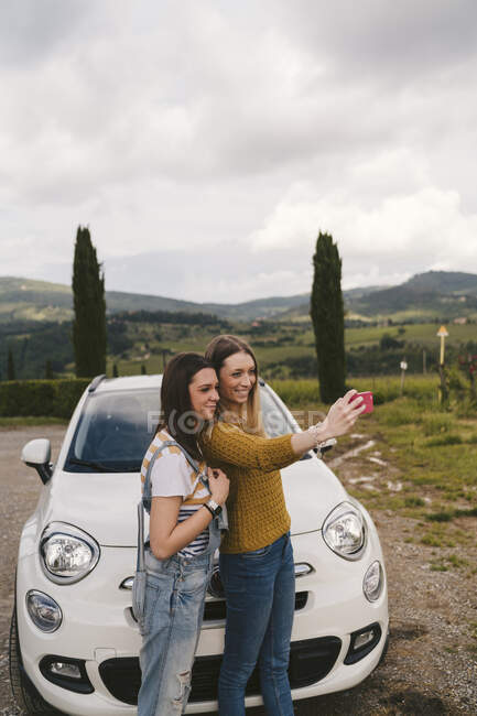 Deux jeunes femmes heureuses debout à côté de la voiture prenant un selfie, Toscane, Italie — Photo de stock