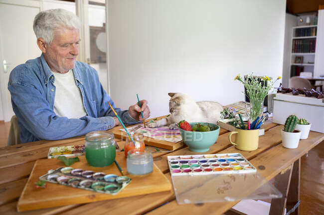Uomo anziano pittura mentre gatto sdraiato sul tavolo a casa — Foto stock