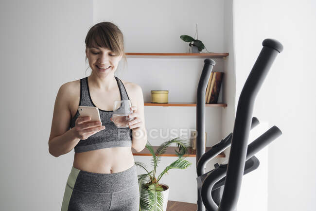 Woman drinking water and using smartphone after performing workout on elliptical trainer at home — Stock Photo