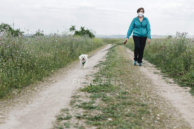 Mulher sênior com máscara protetora vai walkies com seu cão na pista de sujeira — Fotografia de Stock