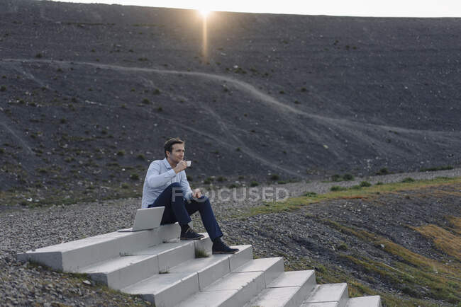 Reifer Geschäftsmann sitzt auf einer Treppe auf einer stillgelegten Grubenspitze neben einem Laptop und macht eine Kaffeepause — Stockfoto
