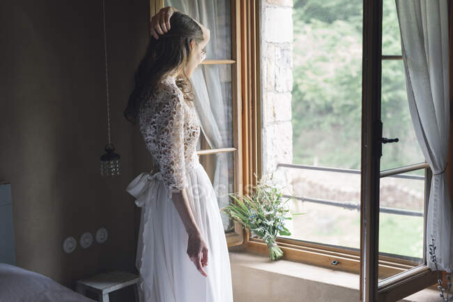 Jeune femme en robe de mariée élégante tenant bouquet regardant par la fenêtre — Photo de stock