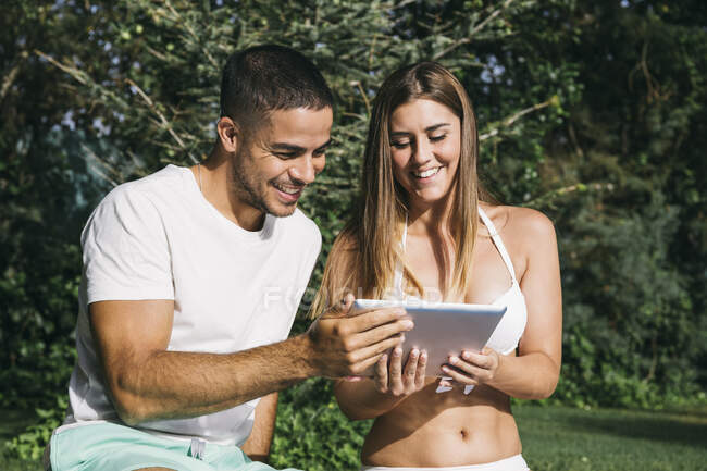 Sorridente giovane coppia utilizzando tablet digitale mentre seduto contro gli alberi a bordo piscina — Foto stock