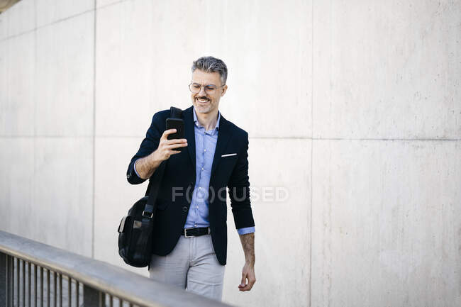 Homme d'affaires souriant aux cheveux gris marchant dans la ville avec un téléphone portable — Photo de stock