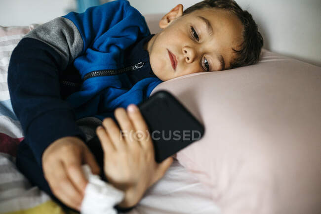 Портрет хворого маленького хлопчика, який лежить у ліжку і дивиться на смартфон. — стокове фото