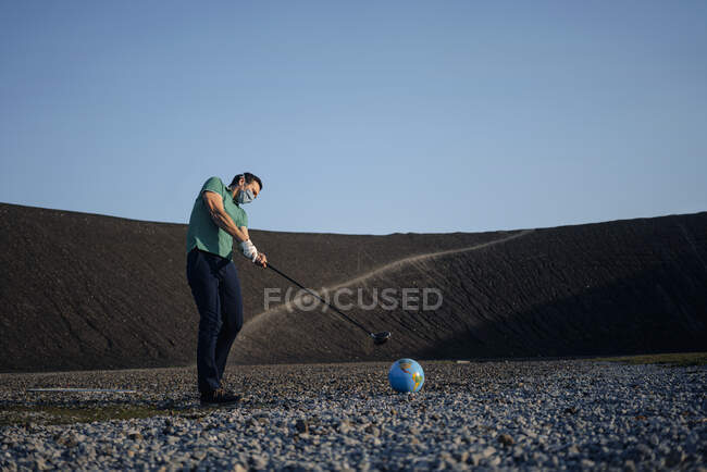 Reifer Mann mit Maske spielt Golf mit einer Erdkugel auf einer stillgelegten Minenspitze — Stockfoto