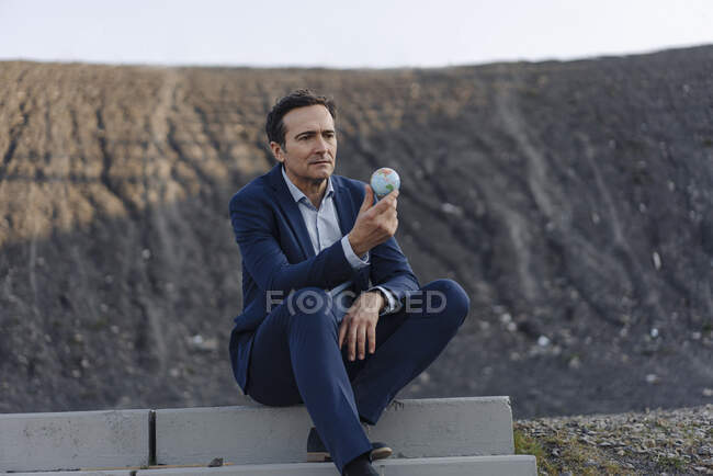 Reifer Geschäftsmann mit einer Miniaturkugel auf einer stillgelegten Minenspitze — Stockfoto
