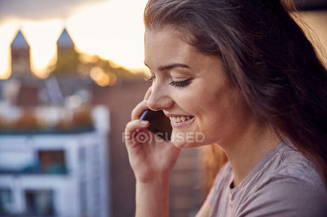 Lächelnde junge Frau am Telefon auf dem Balkon bei Sonnenuntergang — Stockfoto