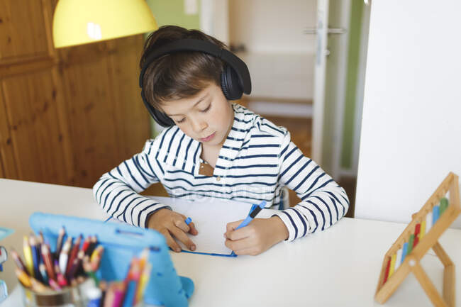 Niño haciendo la educación en casa y la escritura en el portátil, el uso de tabletas y auriculares en casa - foto de stock